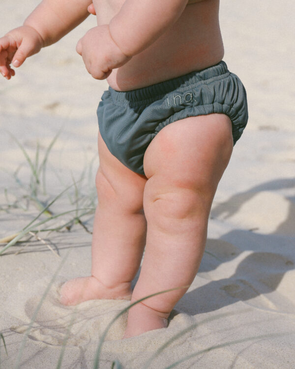 A baby standing in the sand wearing a Lumi Brief Swim Nappy.