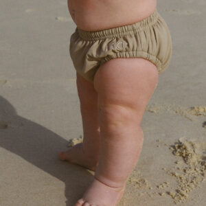 A baby standing on the beach holding a Lumi Brief Swim Nappy.