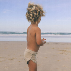A little boy standing on the beach with Mesa Trunks and curly hair.