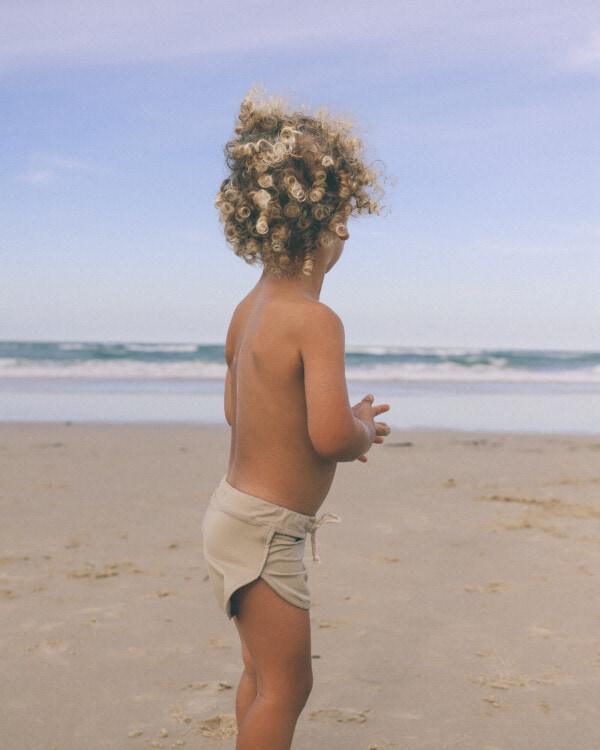 A little boy standing on the beach with Mesa Trunks and curly hair.