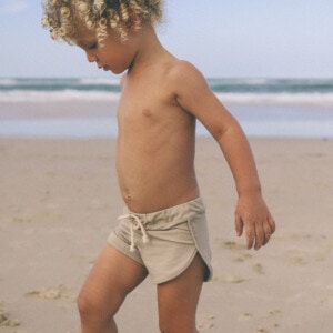 A young boy walking on the beach with Mesa Trunks and curly hair.