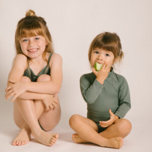 Two little girls sitting on the floor eating the Essentials Range - June One-Piece - Moss Colour.