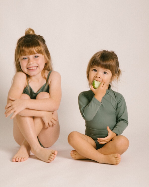 Two little girls sitting on the floor eating the Essentials Range - June One-Piece - Moss Colour.