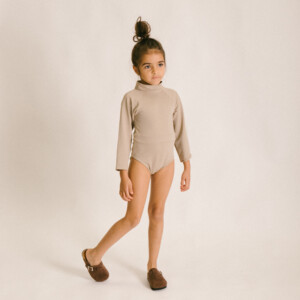 A little girl wearing a June Long Sleeve One-Piece bodysuit and brown slippers.