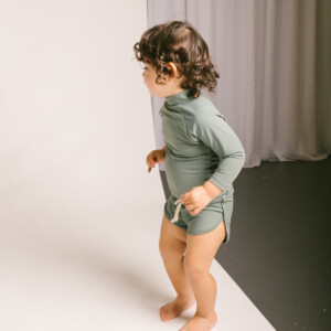 A baby standing on a white background in Mesa Trunks.