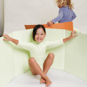 A young girl is sitting on a Playtime Collection - June Long Sleeve One-Piece - Melon.