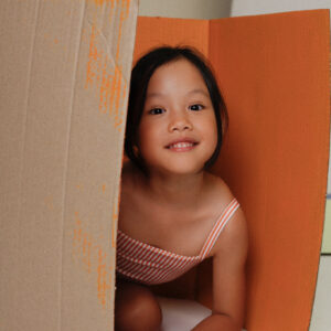 A little girl sitting in the Playtime Collection - Mara One-Piece - Mandarine Stripe.