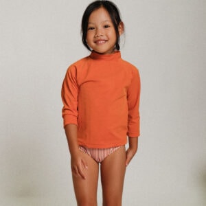 A little girl wearing the Playtime Collection - Nella Rash Shirt in Mandarin and striped shorts.