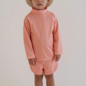 A young boy wearing a Sorbet Summer - Nella Rash Shirt hat and shorts in a coral color.