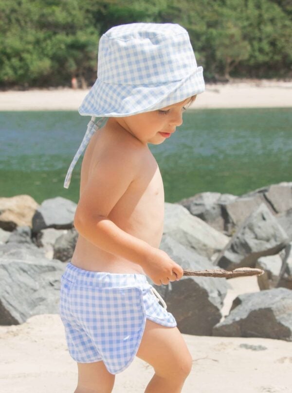 A baby boy wearing Azure and Apricot Gingham - Mesa Trunks on the beach.