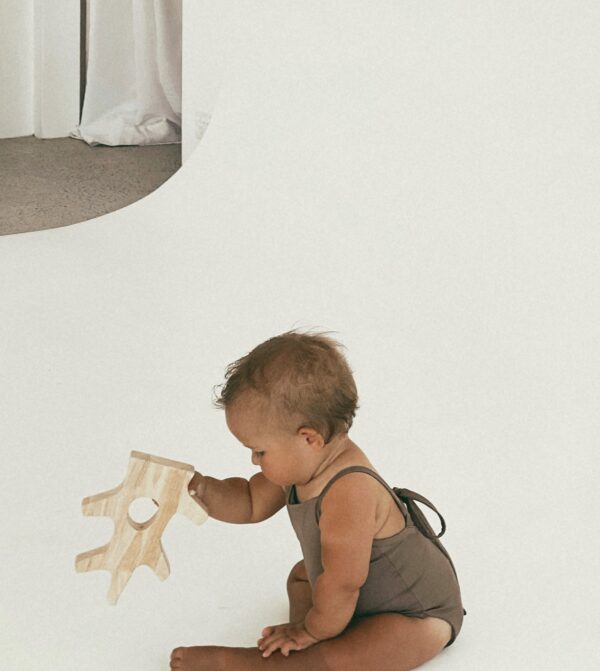 A baby is playing with a wooden toy on the floor wearing the Mara One-Piece.