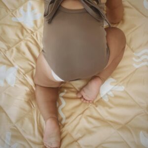 A baby is peacefully sitting on a bed wrapped in a cozy Mara One-Piece.