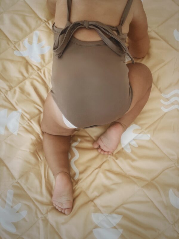A baby is peacefully sitting on a bed wrapped in a cozy Mara One-Piece.