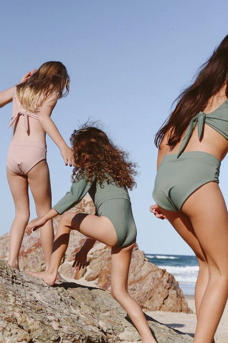 Three girls wearing eco-friendly swimwear in green bikinis, standing on rocks at the beach.