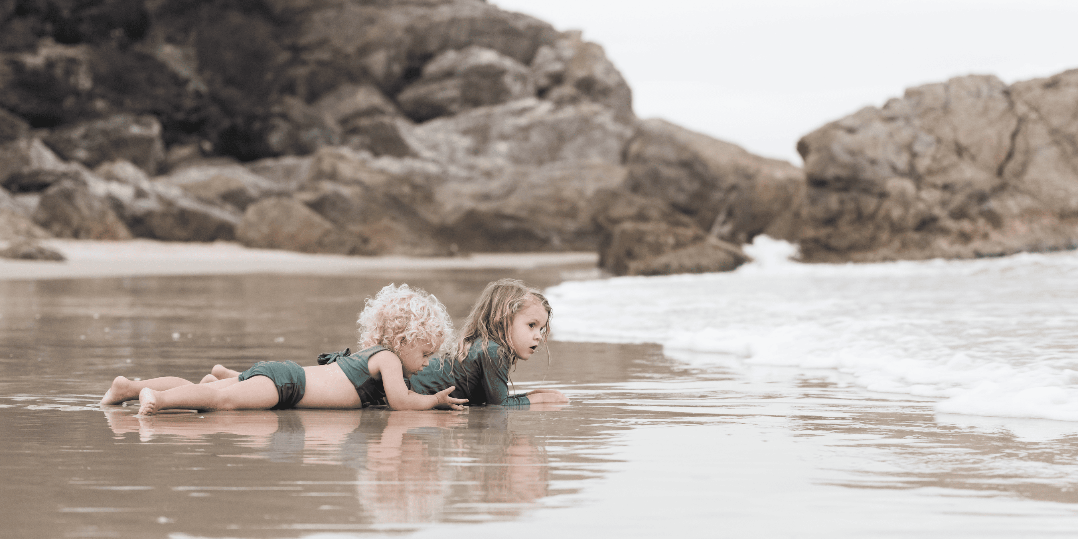 Two eco-friendly children laying on the beach with rocks in the background.