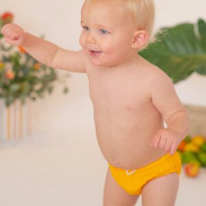 A baby in a Retro Wave By Ina - Lumi Brief Swim Nappy standing in front of a plant.