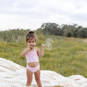 A toddler standing in a field holding a Peach Blossom flower, with a blanket on the ground nearby.