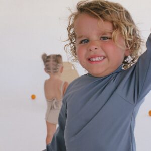 A young boy with curly hair is holding a Essentials Range - Nella Rash Shirt - Mineral Colour.