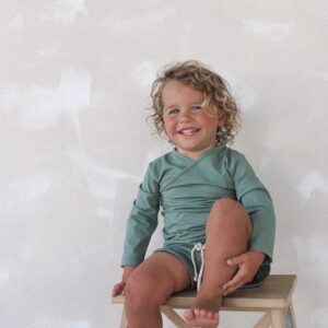 A young child sitting on a stool with a smile on his face, wearing the Essentials Range - Ada Rash Shirt in Moss Colour.