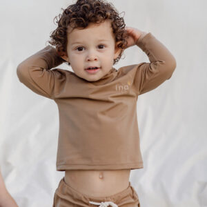 A young child with curly hair dressed in Mesa Trunks - Warm Pecan stands with hands on head against a white backdrop.
