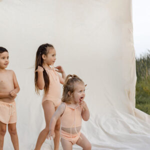 Three children in Luna Bikini - Peach Blossom swimwear playing in front of a plain backdrop outdoors.